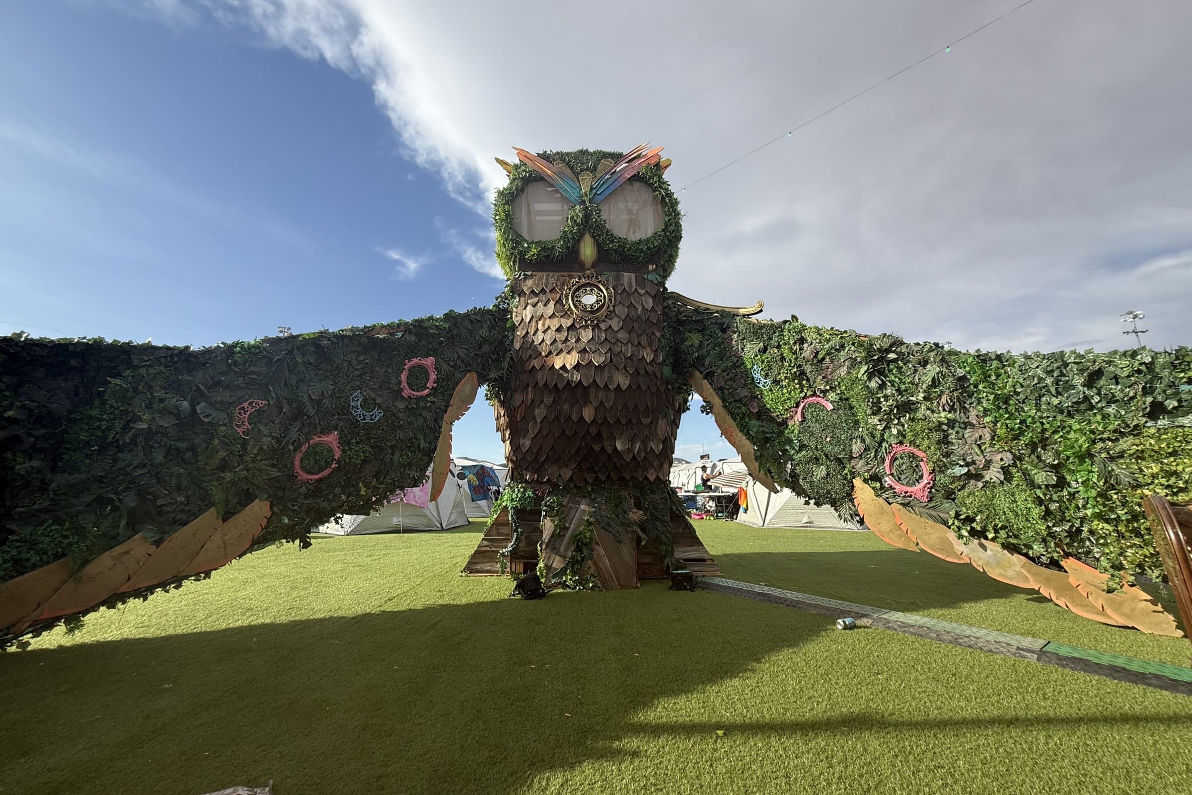 Inside Camp EDC in daylight: the owl installation with its wings spread over the turf, camping pods behind